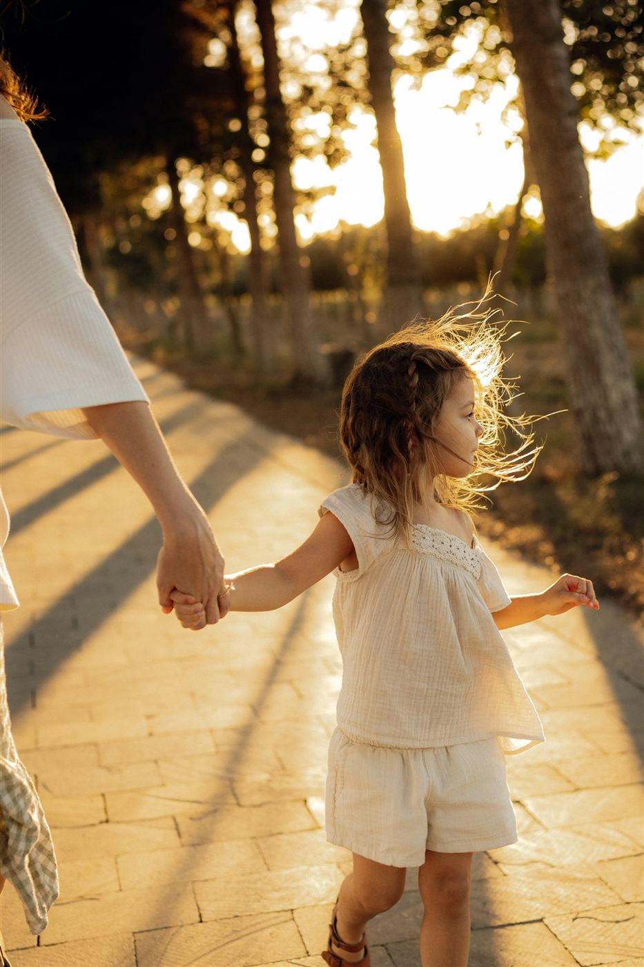 A parent holding a young child's hand on a sunlit path