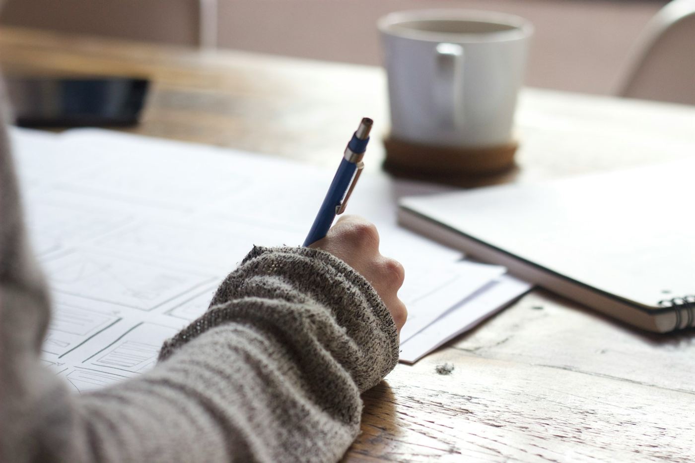 A hand writing notes at a wooden table beside a mug of coffee